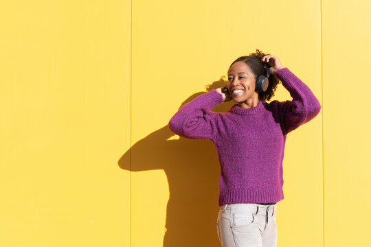 Happy woman wearing wireless headphones listening to music in front of yellow wall
