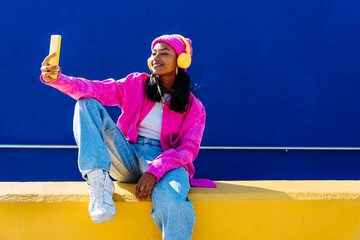 Smiling woman taking selfie through smart phone sitting on wall