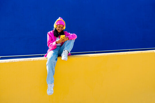 Smiling young woman text messaging through smart phone sitting on wall