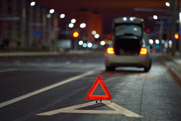 Broken down car on city street at night, red triangle - emergency stop sign behind broken blurred...
