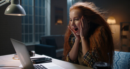 Positively surprised redhead caucasian girl looking at laptop, wing blowing her hair. Model reading...