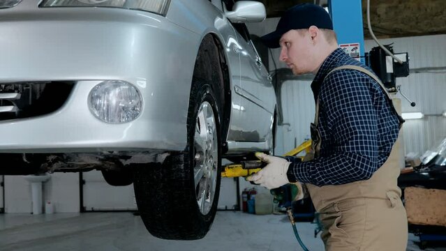 The mechanic removes the wheel of the car. A young auto mechanic in uniform works in a car service with a raised car