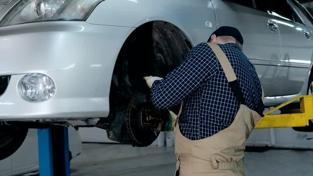 An auto mechanic inspects the braking system of a car. A mechanic in uniform works in a car service with a raised car