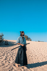 A Japanese warrior in a traditional kimano armed with a katana sword on a sandy shore next to the ocean during sunset. A man from medieval Asia. Reconstruction of cultural heritage. Culture in Japan. 