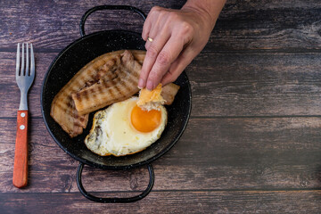 sarten de huevos con panceta encima de una mesa de madera, mujer mojando pan en la yema del huevo