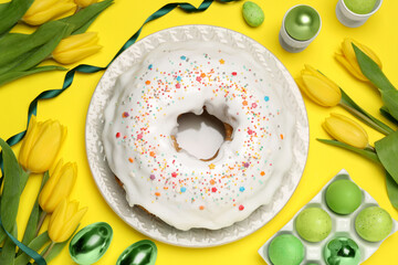 Easter cake with sprinkles, painted eggs and tulips on yellow background, flat lay