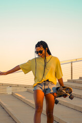 Young pretty brown-skinned woman with long black braids having fun holding a skateboard with her hand dressed in warm colored summer clothes standing on a concrete stairs on the seafront in summer.