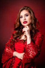 Portrait of a young, attractive vampire woman in a red rococo dress posing isolated against a dark background with red backlights.