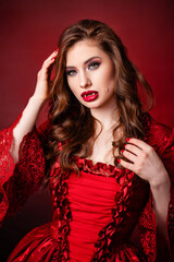 Portrait of a young, attractive vampire woman in a red rococo dress posing isolated against a dark background with red backlights.