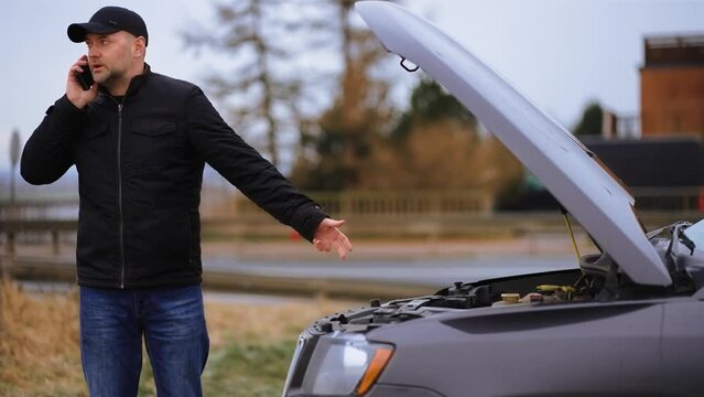 Sad man standing near broken car with opened hood and speak by mobile phone to rescue mechanic. Stressed male on road with problem emergency insurance auto