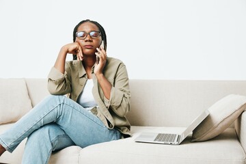Fototapeta na wymiar Stylish African American woman business freelancer working sitting on the couch at home in a laptop and phone, business calls and correspondence, home clothes and eyeglasses, light interior background