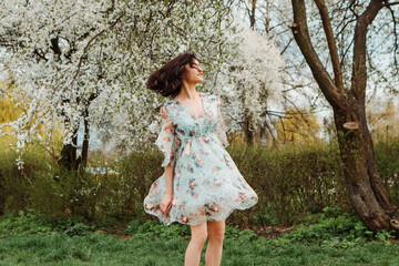 Portrait of charming pretty woman dressed flowery dress spinning around having fun laughing smiling near apple cherry tree blossoms blooming flowers in the garden park in early spring nature

