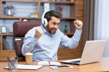 Obraz premium Mature businessman in shirt and headphones watching sports match at workplace inside office, man cheering having fun at work, watching video on laptop.