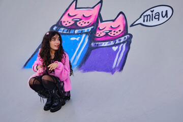 Teenage girl dressed in gothic style posing squatting on a skateboarding rink with graffiti in background