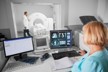 Medical computed tomography or MRI scanner. Back view of blonde nurse sitting at computer, doctor...