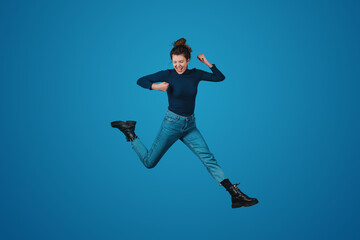 Naklejka na ściany i meble Full length view of joyful woman jumping expressing positive emotions isolated over blue background. Positive person. People lifestyle portrait. Attractive