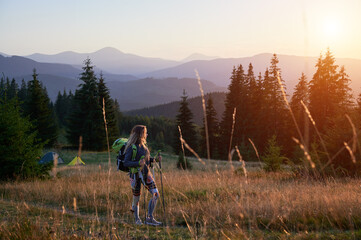 Woman traveller hiking outdoors at sunset. Sporty, slim woman traveling in mountains. Female tourist carrying backpack, using trekking sticks, admiring landscape. Concept of harmony with nature. - obrazy, fototapety, plakaty