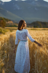 Beautiful girl in long white dress on the wheat field enjoying golden sunset outdoors. Her hand touching of spikes
