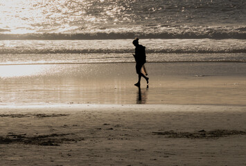 Person in silhouette, walking along beach during golden sunset, with cell phone, beach combing with collection bag. 