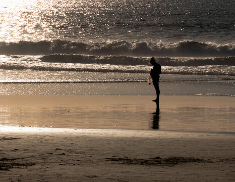 Person in silhouette, walking along beach during golden sunset, with cell phone, beach combing with collection bag. 