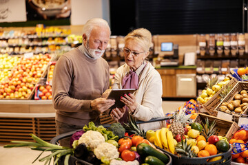 An old couple is looking at the groceries list on tablet at the supermarket.