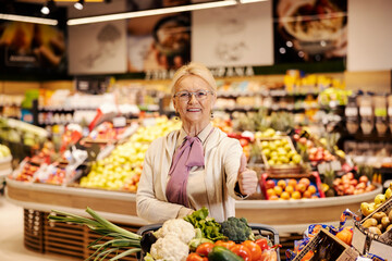 A senior woman is purchasing at the supermarket and giving thumbs up while smiling at the camera.