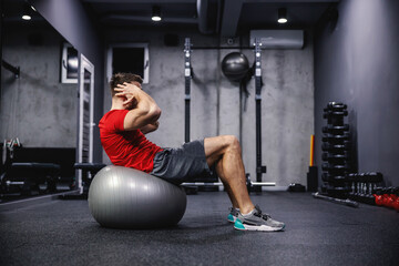 Fototapeta na wymiar Exercises for the abdomen and whole body, a good balance. A man in excellent shape does sit-ups on a fitness ball indoors at the gym. Personal training and sports motivation. Healthy and strong body