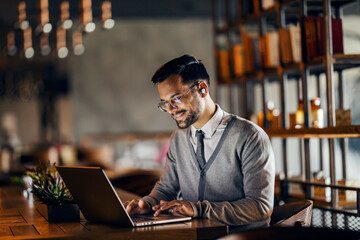 A happy young trendy businessman is sitting in cafeteria and working remotely on the laptop.