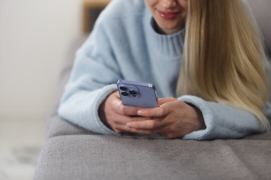 Young woman lying on couch and typing a message on a smart phone. Close up photo of white female person using modern mobile phone for communication