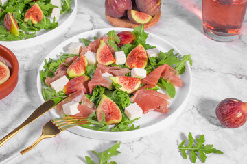 Autumn figs salad with arugula, feta, raw ham in white plate on light marble background served with rose wine. Flexitarian diet, paleo diet fall or summer salad, close up