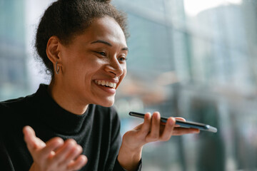 Smiling woman recording audio message on mobile phone while while sitting in cozy cafe near window