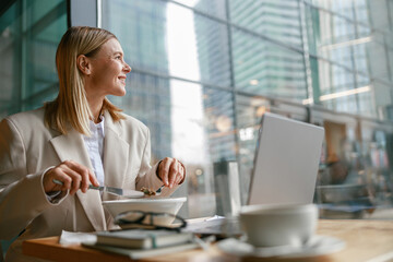 Smiling businesswoman is having a business lunch and working on laptop in cafe. Blurred background