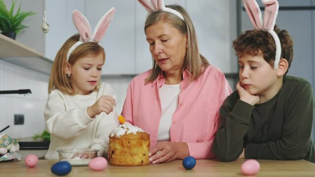 Happy Grandmother with Grandchildren Wearing Bunny Ears Decorating Easter Cake at Home Together. Family Spending Time Together. Preparing for the Easter Holiday