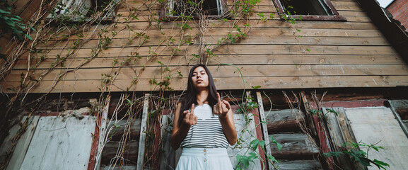 Crazy girl shows middle finger. Beautiful mad country girl near old rustic house with broken...