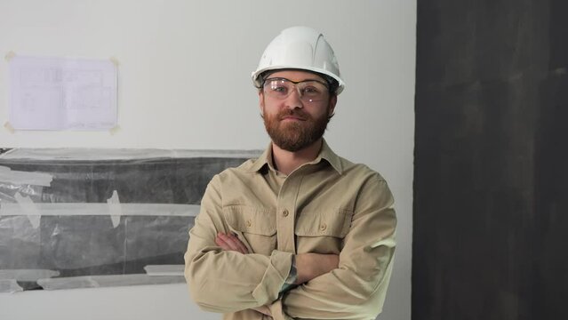 Portrait of a young bearded man in a white helmet, goggles and beige shirt folding his arms on his chest. Professional architect engineer or foreman looks into the camera while on a construction site