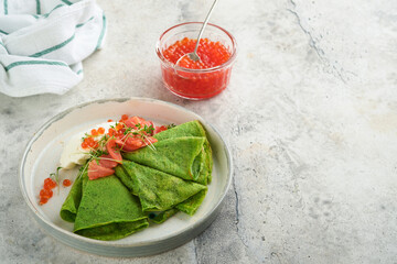 Green pancakes. Round thin green spinach crepe pancakes with red and black caviar and yogurt sauce on gray concrete background. Healthy vegan breakfast. National Pancake Day or Fat Tuesday Day.