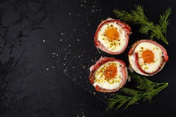 baked chicken eggs with bacon greens and spices on a stone board on a black textured background. rustic still life with copy space. top view