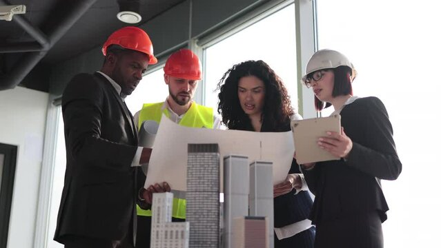 Happy multiracial architects, engineers and designers standing near skyscrapers buildings maquette during meeting at boardroom. Men and women colleagues leading meeting for architectural project.