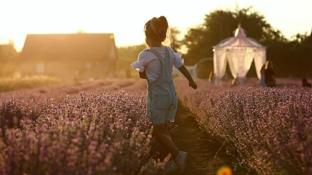 Adorable happy child girl is having fun and jumping in lavender field on summer warm day. Hyperactive smiling little kid in sunglasses and jeans jumpsuit on nature. Happy childhood concept.