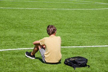 Tired sad alone teenage boy with backpack sitting in empty sport stadium outdoors. Back view