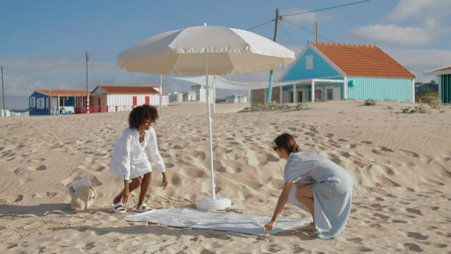 Girlfriends Spread Picnic Blanket On Sandy Beach. Two Girls Enjoying Summer Day