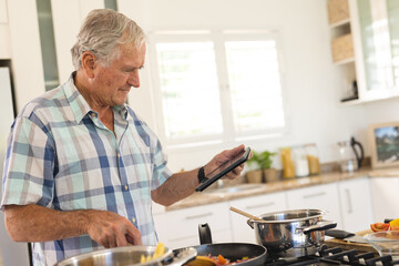Happy senior caucasian man cooking dinner and using tablet in kitchen