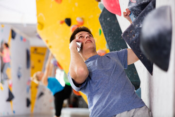 European young man having telephone conversation while grabbing ledges of artificial climbing wall in bouldering centre.