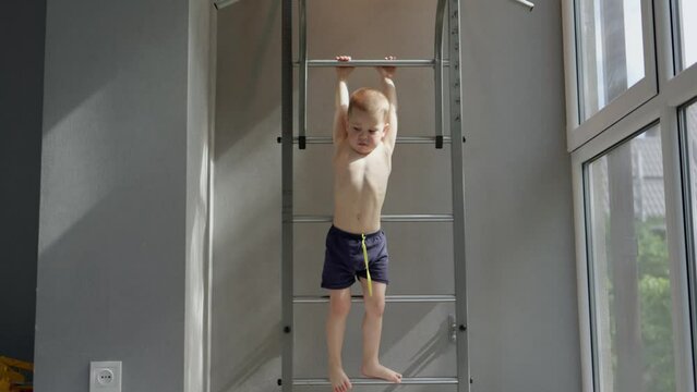little kid boy hanging on gymnastics wall bars. male child climbing wall ladder swedish wall at home. kid holding hands bar, jumping running away camera. sunny day indoor room. healthy child concept