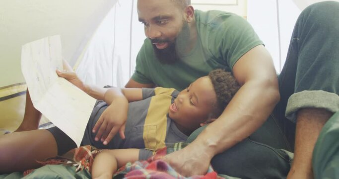 Happy african american father and son sitting in tent and reading, in slow motion