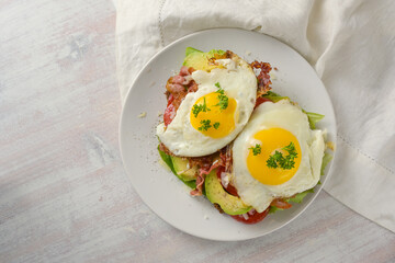 Brunch sandwich with fried egg, avocado and bacon on a white plate and a light wooden table, high angle view from above, copy space