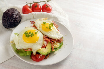 Avocado and fried egg and with bacon, tomato and lettuce on a white plate and a light wooden table, delicious brunch sandwich, copy space, selected focus