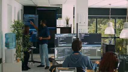 Diverse team of software engineers analyzing code on wall screen tv, using digital tablet and...