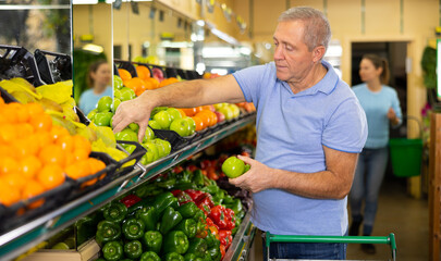 in modern supermarket, elderly man carefully selects apples and puts them in shopping cart