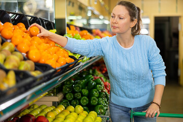 Glad middle-aged woman customer buying fresh mandarins at the counter in big greengrocery